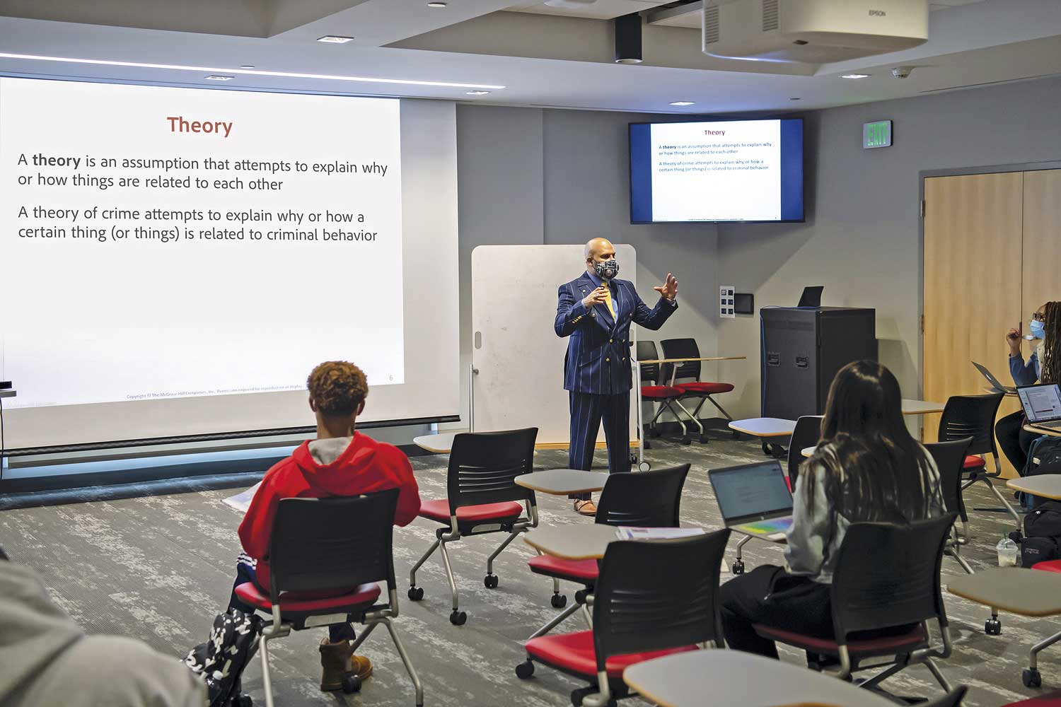 Seen here undivided, the Divisible Classroom accommodates a large, socially distanced class taking a Criminal Justice course. A TLP Pro desktop touchpanel is on top of the AV equipment rack positioned against the wall below the exit sign. This touchpanel augments wall mounted touchpanels on either side of the divisible room, making it convenient to operate both sides of the room from a single location when necessary.